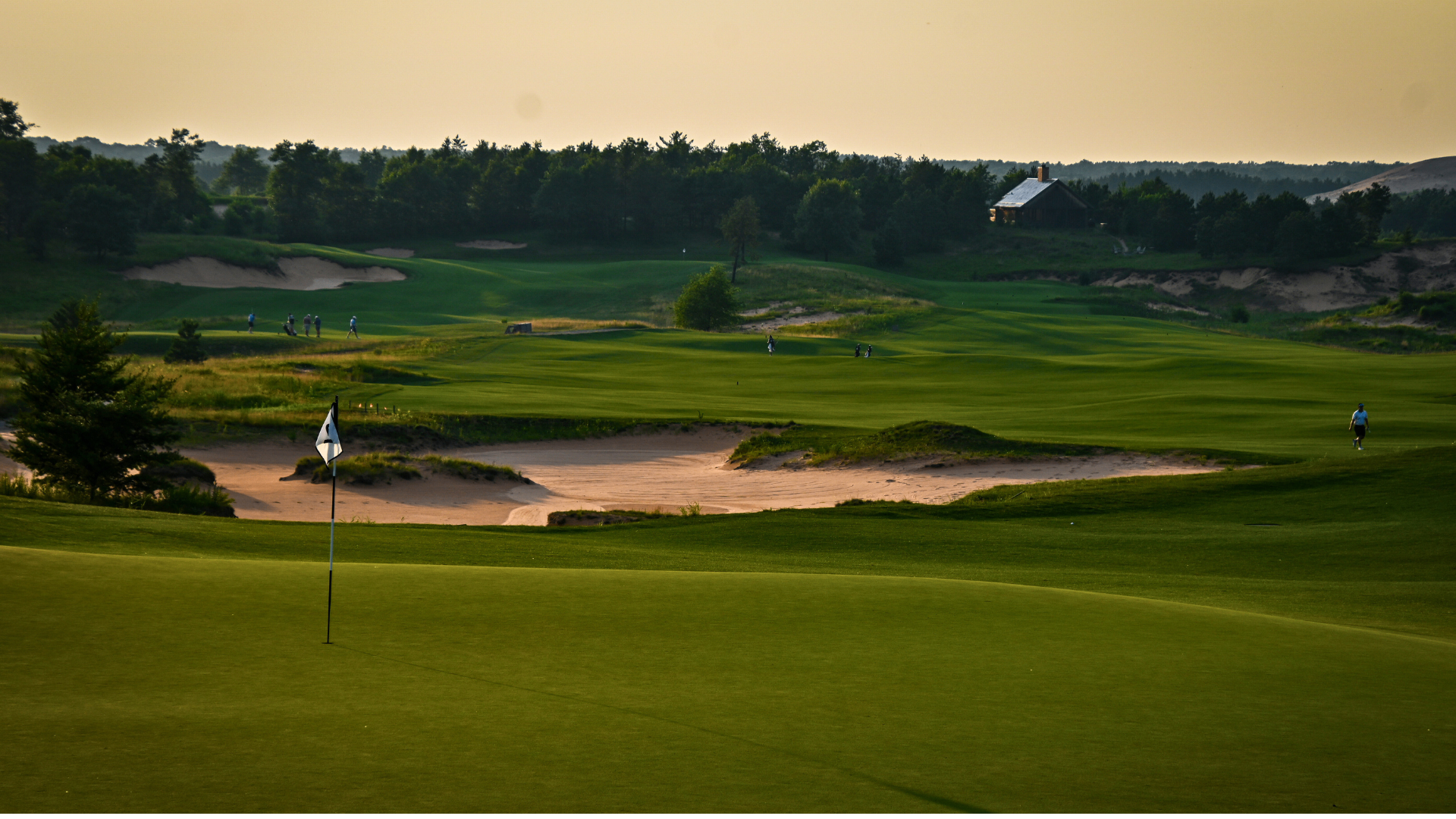 Sandy links-style course at golden hour — green, flag, and expansive waste bunker with golfers in the distance